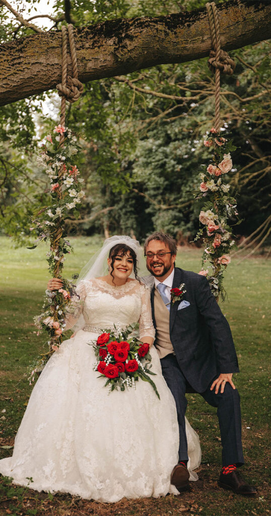 A bride in a white lace gown and veil, holding red roses, sits on a floral-decorated swing beside a groom in a navy suit and beige vest. Captured by a talented photographer at Bardney Hall, they smile amid the lush park setting, surrounded by trees and floral garlands on the swing's ropes. © Aimee Lince Photography