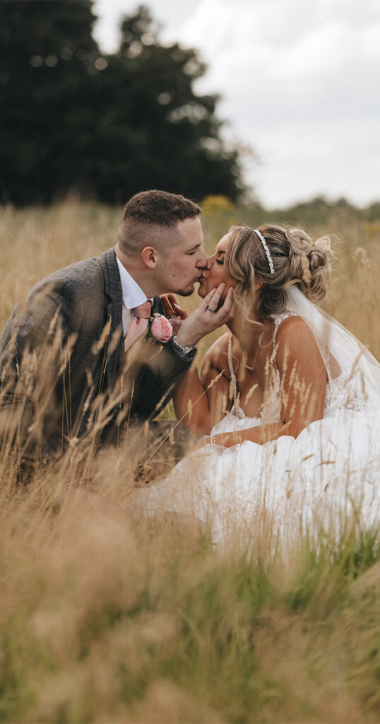 In a stunning display of wedding photography, a couple kisses in a field of tall grass. The groom sports a dark suit with a pink tie and boutonniere, while the bride radiates elegance in her white dress, veil, and tiara. They sit amid lush greenery, blurred trees behind them under the cloudy sky near Oaklands Hall Hotel. © Aimee Lince Photography