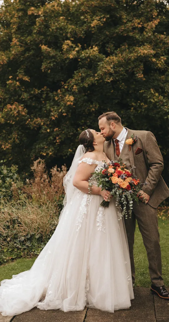 Photography captures a bride and groom sharing a kiss beneath the vibrant hues of autumn. The bride, in her white lace-adorned gown, holds a bouquet of orange, red, and peach flowers. The groom complements her in a brown suit with a rust-colored tie as they stand amidst lush greenery on a stone path. © Aimee Lince Photography - Wedding photographer in Lincolnshire, Yorkshire & Nottinghamshire