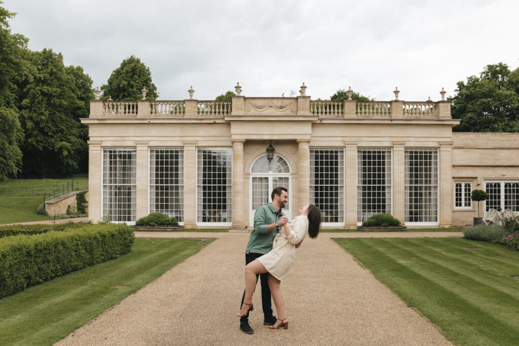 A couple dances in front of a grand building with large glass windows and ornate stonework at Rushton Hall in Kettering. The man, in a green shirt and black pants, dips the woman who is wearing a beige dress and heels. The setting includes well-manicured lawns, bushes, and a gravel path leading to the building.</p>
<p>. © Aimee Lince Photography