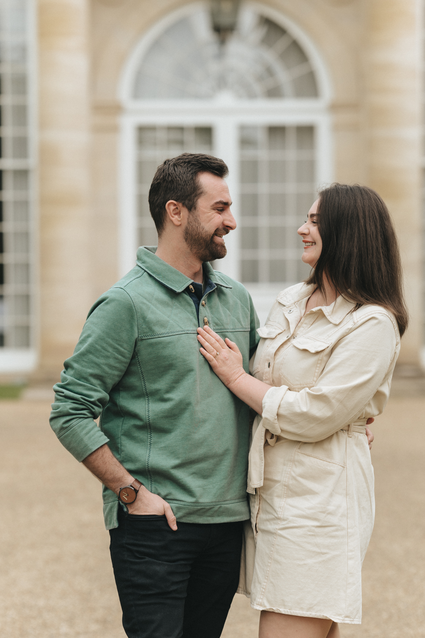 A man and a woman stand close, smiling at each other. The man, with short dark hair and a beard, wears a green sweatshirt and black pants. The woman, with shoulder-length dark hair, wears a light beige dress with long sleeves. They stand in front of a building with large arched windows. © Aimee Lince Photography