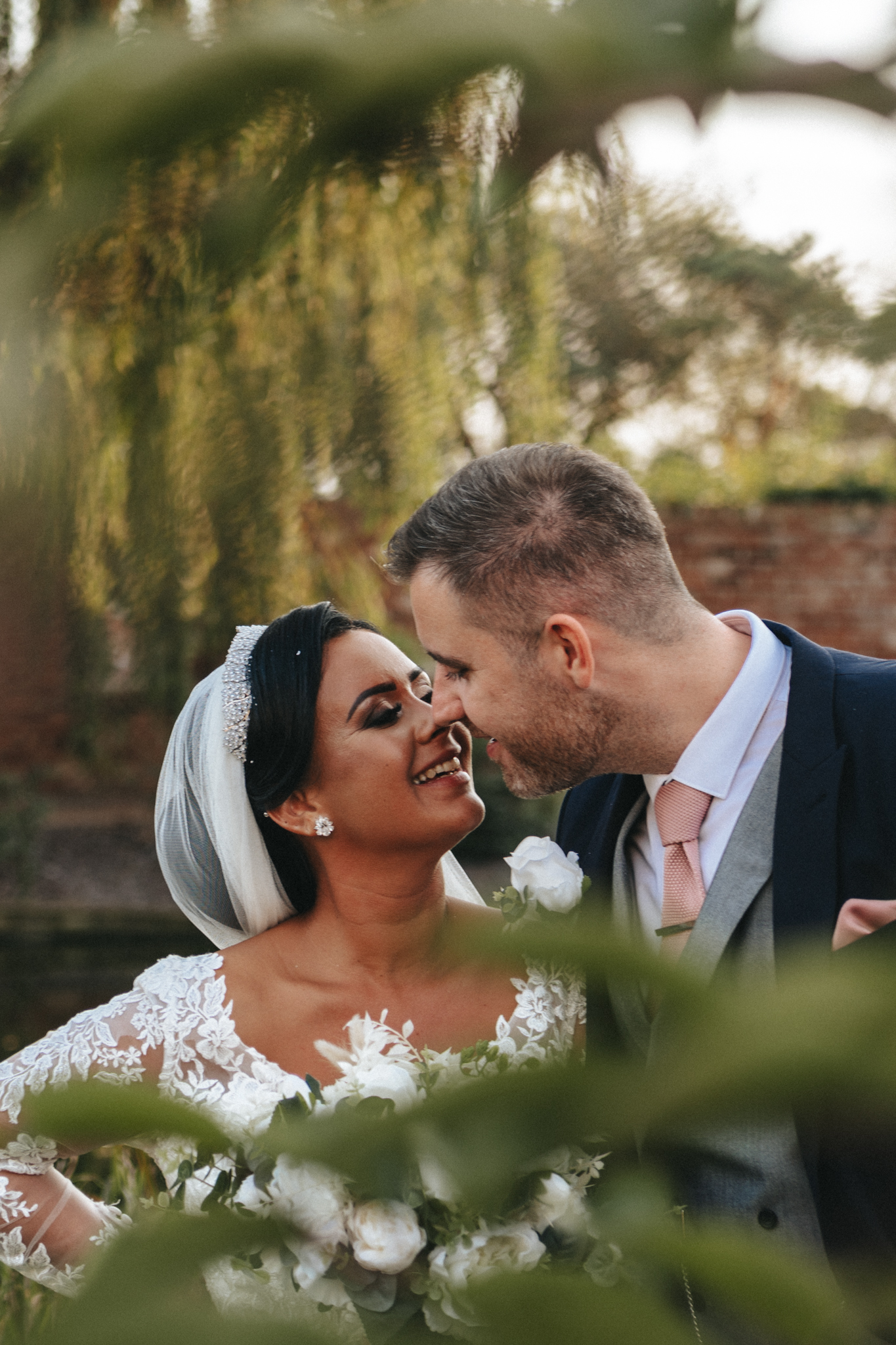 A bride and groom smile at each other, their foreheads touching. The bride is wearing a white lace gown and a veil, holding a bouquet of white roses. The groom is dressed in a dark suit with a pink tie. Green foliage frames the couple, with an outdoor background of trees and a brick wall behind them. © Aimee Lince Photography