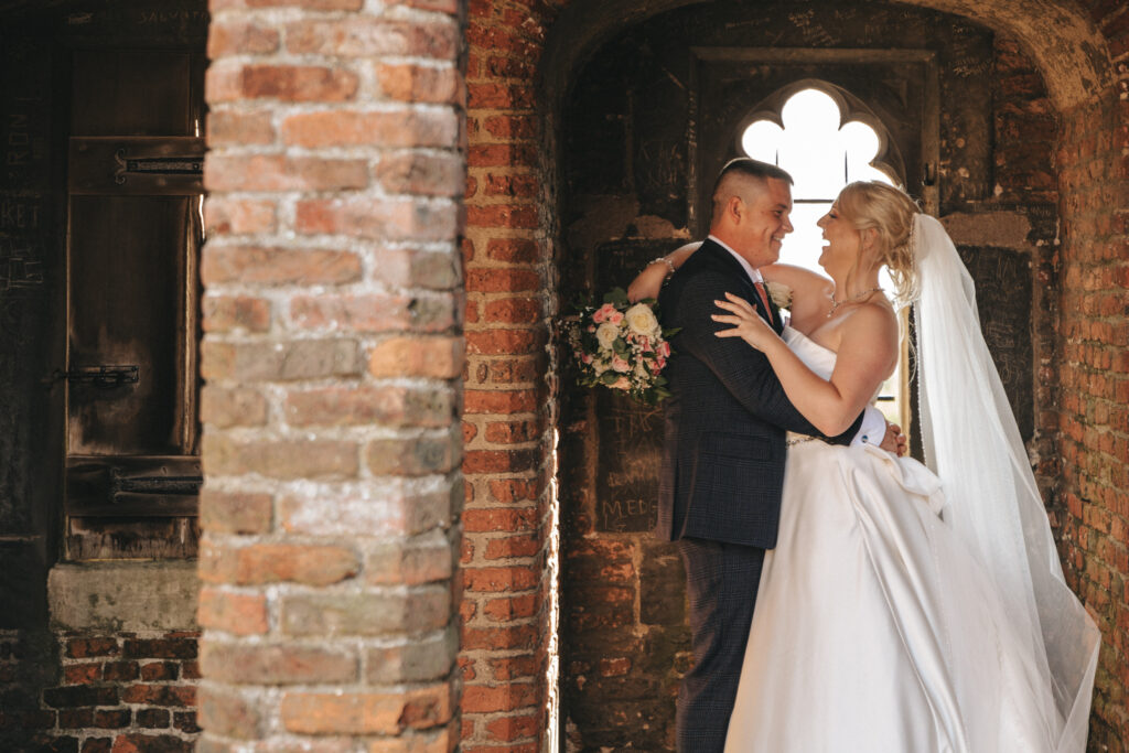 A newlywed couple stands in an intimate embrace within a rustic, brick archway at Tattershall Castle. The bride, in a white gown and veil, holds a bouquet of flowers. The groom, in a dark suit, smiles tenderly at her. Soft sunlight filters through a clover-shaped window, illuminating the romantic moment. © Aimee Lince Photography