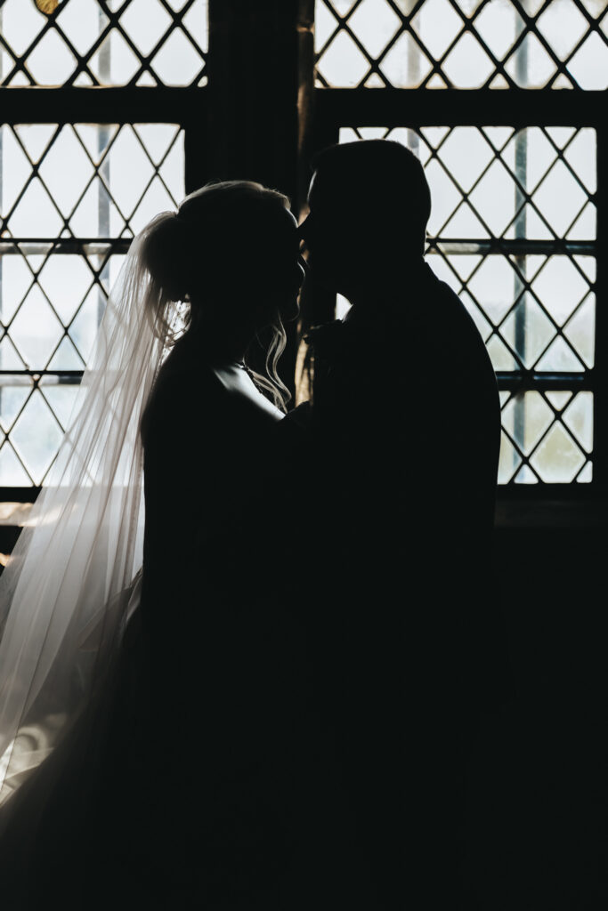 Silhouetted couple sharing a tender moment in front of a large window with a diamond lattice pattern. The backlighting creates a striking contrast, highlighting the bride's veil and the groom's profile, adding a romantic and dreamy atmosphere. © Aimee Lince Photography