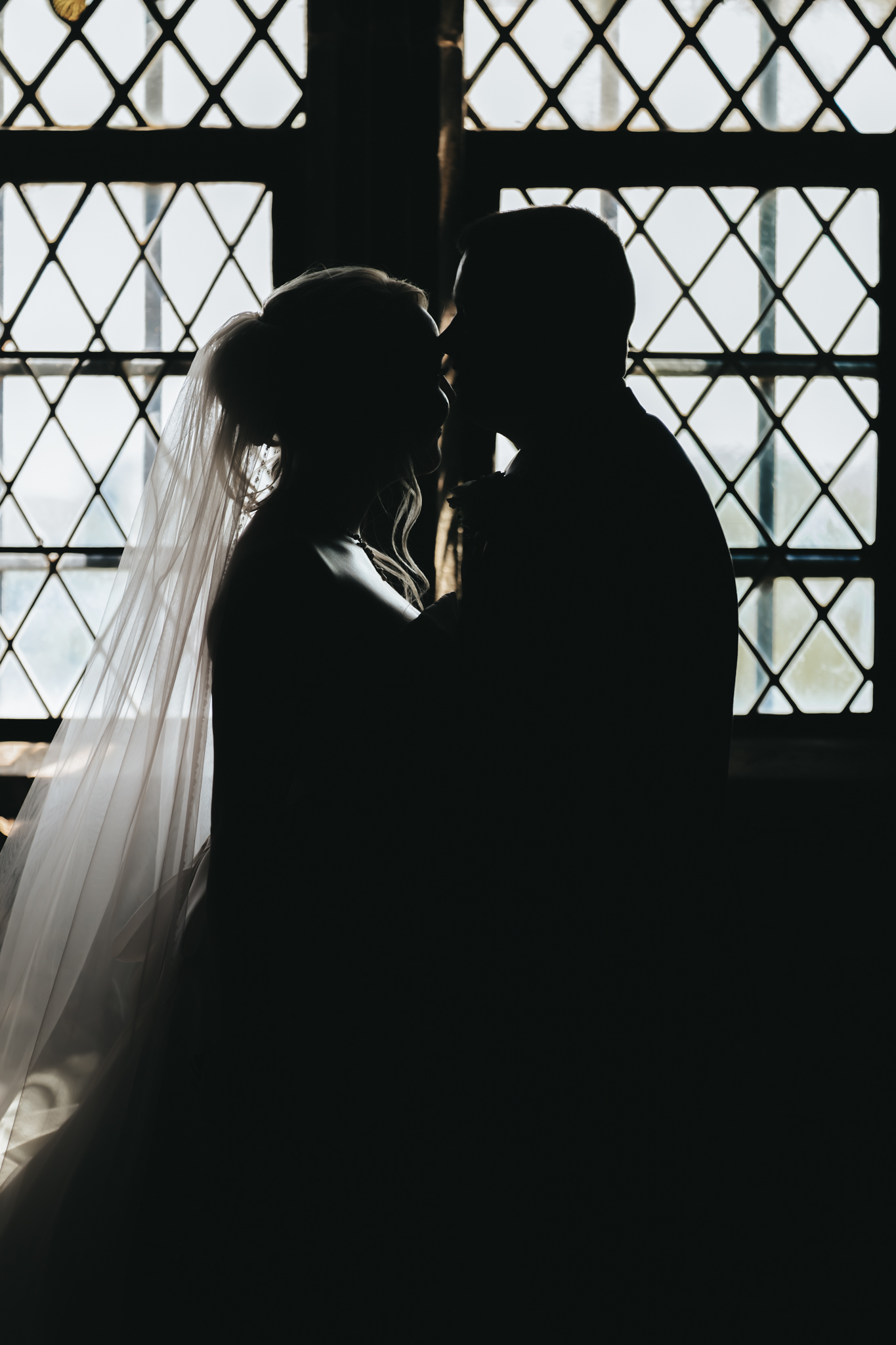 Silhouetted couple sharing a tender moment in front of a large window with a diamond lattice pattern. The backlighting creates a striking contrast, highlighting the bride's veil and the groom's profile, adding a romantic and dreamy atmosphere. © Aimee Lince Photography