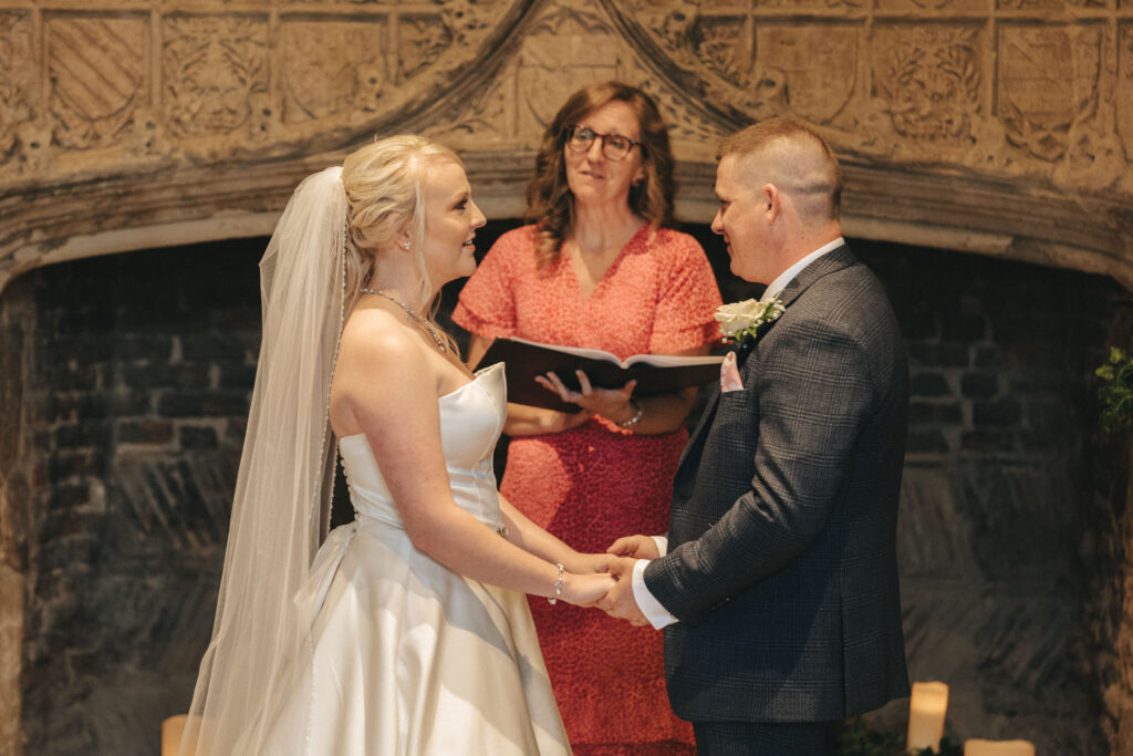 A bride in a white gown and veil holds hands with a groom in a gray suit, framed by the ornate stone fireplace of Tattershall Castle. Behind them, a woman in a pink dress officiates the ceremony with an open book. Captured by an adept photographer, the intimate Lincolnshire setting is warmly lit. © Aimee Lince Photography