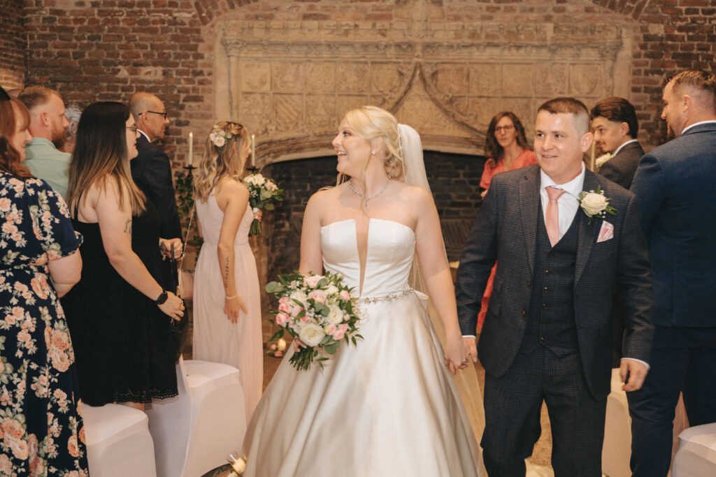 A bride in a satin gown and veil holding a bouquet, and a groom in a dark suit with a pink tie, walk down the aisle smiling. Guests in formal attire, some applauding, are seated on either side. The Nottinghamshire setting boasts rustic brick walls and charming stone architectural details.
. © Aimee Lince Photography