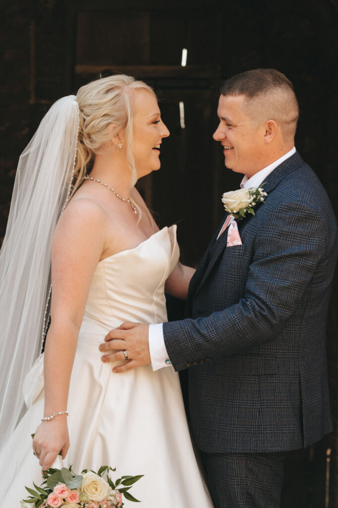 A bride in a strapless white gown and veil holds a bouquet of pink and white roses, smiling at the groom. The groom, wearing a dark checkered suit with a pink tie and boutonniere, gazes at her lovingly. Captured beautifully by wedding photography against Tattershall Castle's blurred backdrop. © Aimee Lince Photography