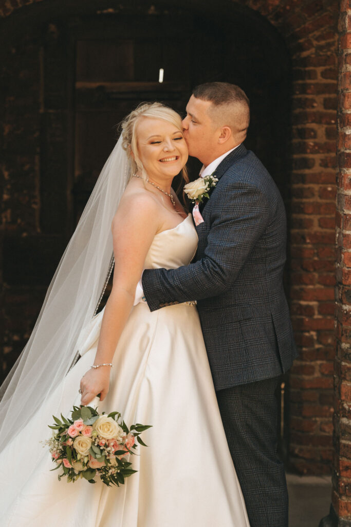A bride in a white gown smiles with her bouquet of pink and white flowers as a groom in a blue patterned suit kisses her cheek. Standing before a rustic brick wall, the couple embodies pure joy on their wedding day at Tattershall Castle in Nottinghamshire. © Aimee Lince Photography