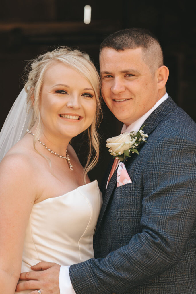 A bride and groom smiling and posing together in Nottinghamshire. The bride wears a strapless white dress, veil, and a pearl necklace, with her hair styled elegantly. The groom is in a blue checkered suit with a white rose boutonniere and a pink pocket square. Captured by their photographer, they stand closely, looking happy. © Aimee Lince Photography