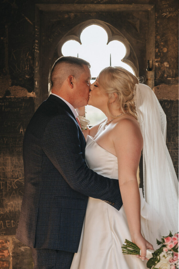 A couple in wedding attire shares a kiss in front of a gothic-style window, captured beautifully by their photographer. The groom wears a dark plaid suit and the bride, in a strapless white gown with a veil, holds pink and white flowers. The stone wall is etched with names in Nottinghamshire. © Aimee Lince Photography