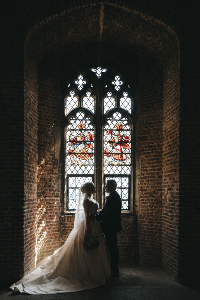 A bride and groom stand silhouetted in front of a large, ornate stained glass window within a brick-walled interior reminiscent of Tattershall Castle. The bride wears a flowing gown and veil, holding a bouquet, while the groom is in a dark suit. Soft light filters through the window, casting a warm glow. © Aimee Lince Photography