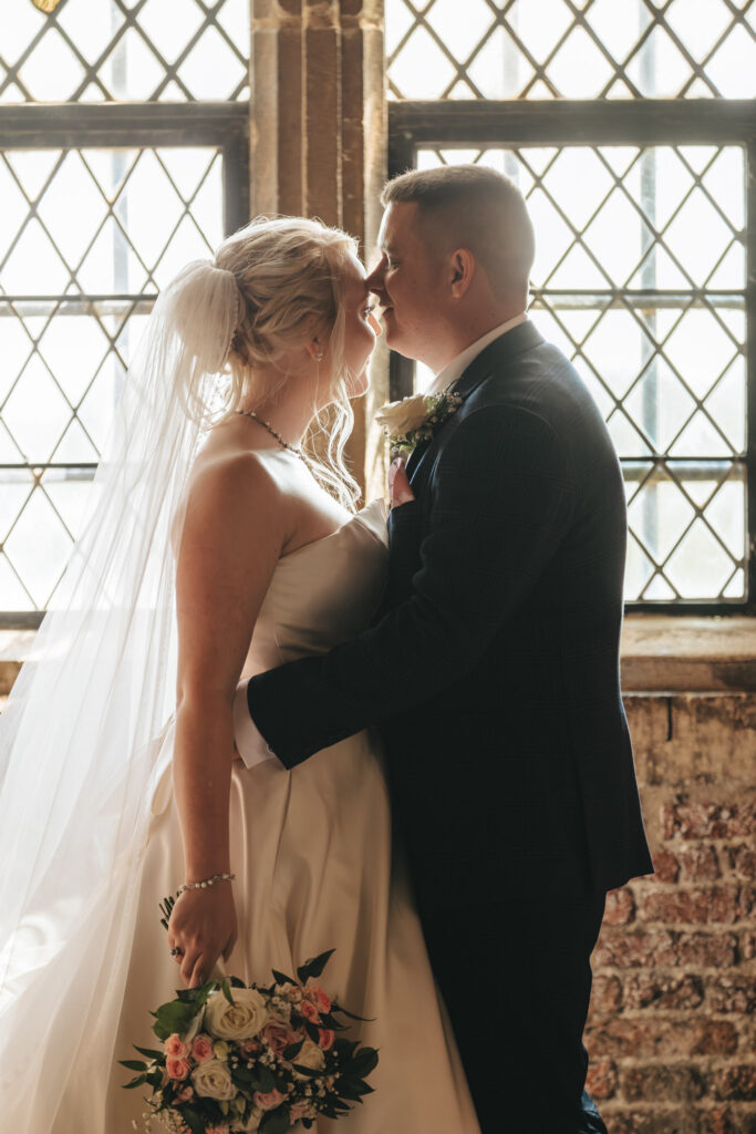 A bride and groom stand facing each other in front of a large, sunlit window with diamond-patterned panes at Tattershall Castle. The bride holds a bouquet of pink and white flowers, wearing a flowing gown and veil. A photographer captures their tender embrace as the groom, in a dark suit, leans in for a kiss. © Aimee Lince Photography