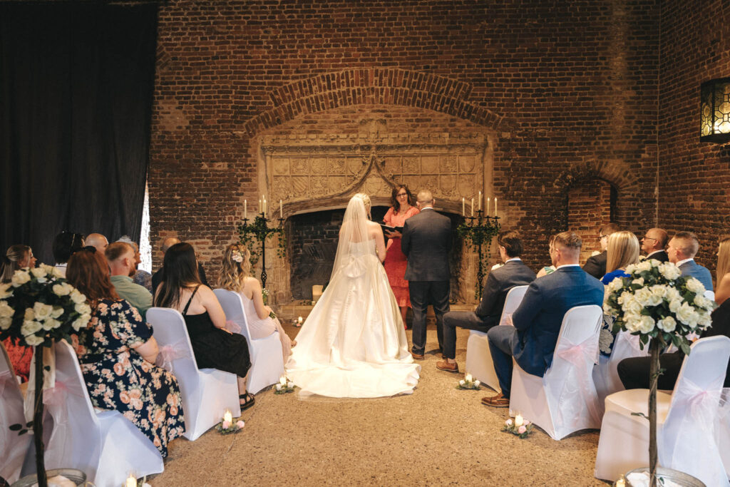 A wedding ceremony in a brick-walled venue unfolds beautifully, like a perfect scene for photography. A bride in a white gown stands facing the priest with her groom beside her, as guests sit on flower-adorned chairs. Tattershall Castle adds an air of Nottinghamshire elegance to the occasion. © Aimee Lince Photography