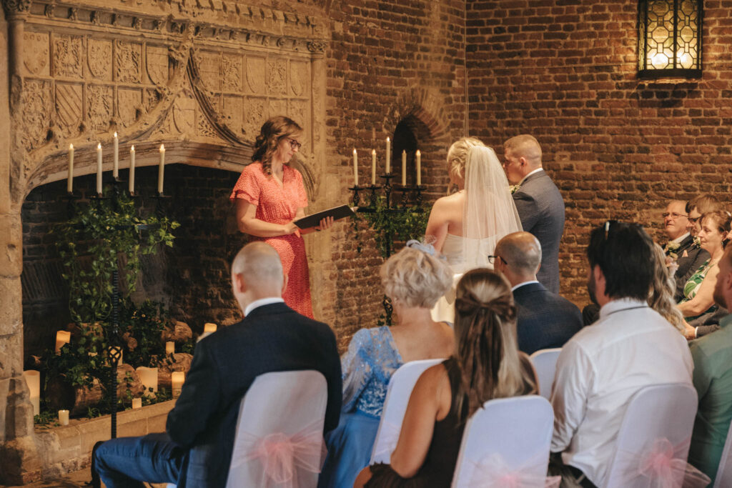 A wedding ceremony unfolds in a rustic brick venue reminiscent of Tattershall Castle, with a large fireplace adorned with candles and greenery. A woman in a pink dress officiates beside the couple, as guests sit on chairs with white covers and pink bows. An ornate lantern hangs above. © Aimee Lince Photography