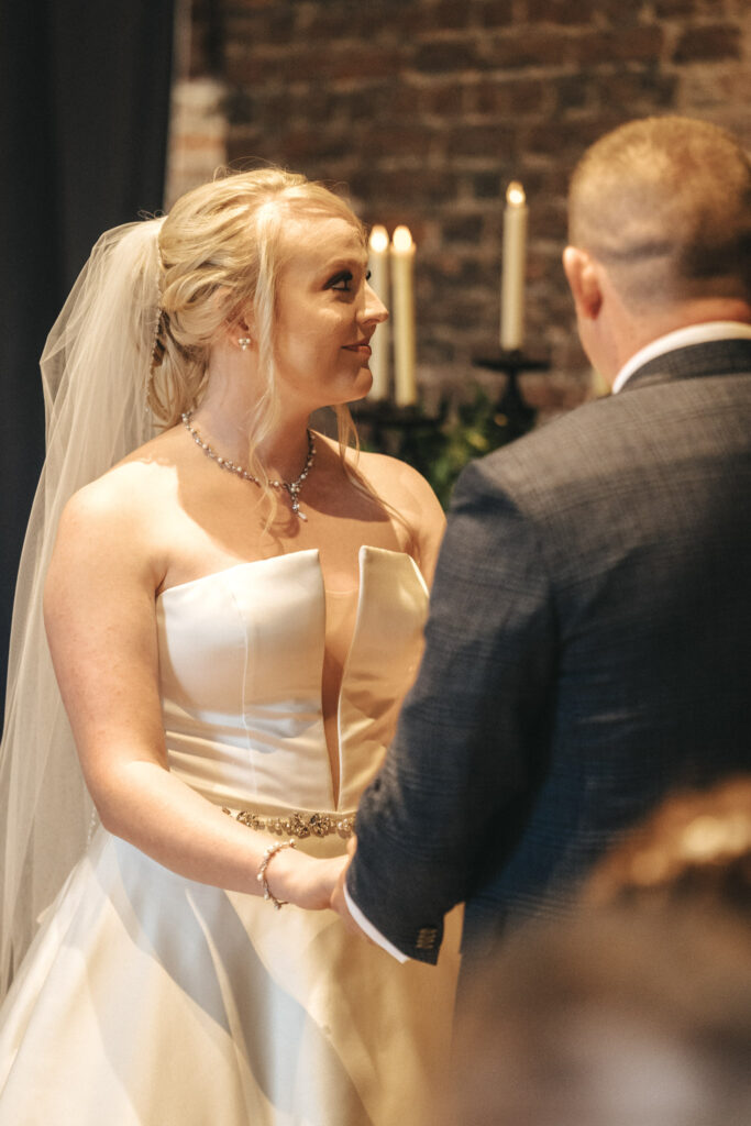 The bride and groom, captured by a talented photographer, are holding hands indoors. She glows in her white dress and veil as he stands in a dark suit. Behind them, lit candles against the brick wall create a warm, intimate atmosphere—a perfect moment of love captured in Lincolnshire. © Aimee Lince Photography