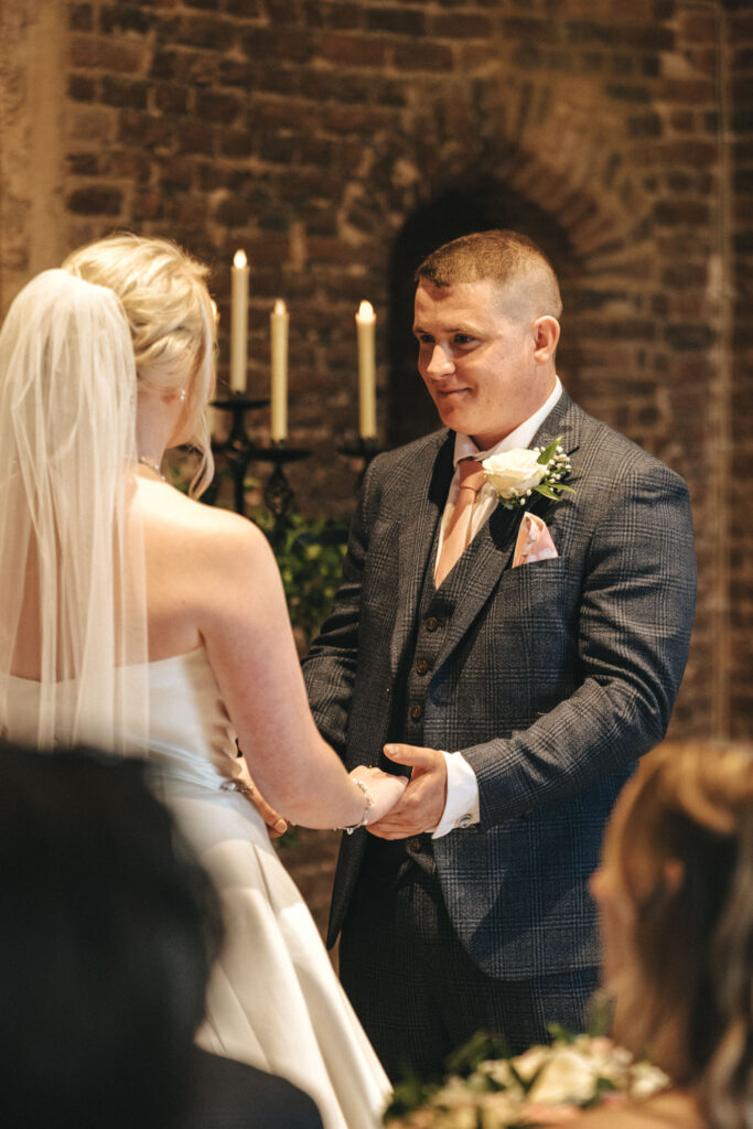 A couple stands facing each other during their wedding ceremony at Tattershall Castle. The bride, in a strapless white gown and veil, holds hands with the groom in a blue checkered suit. Lit candles highlight the rustic brick walls, beautifully captured by their Nottinghamshire photographer. © Aimee Lince Photography