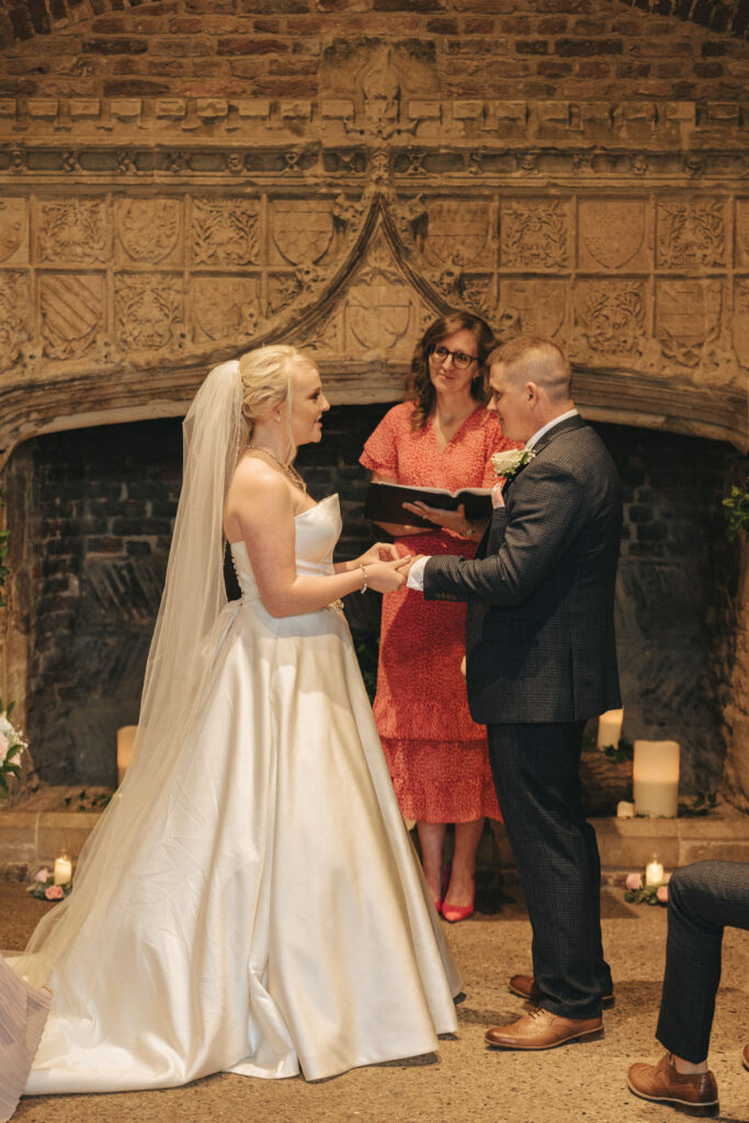 A bride and groom stand holding hands during their wedding ceremony in front of an ornate, carved stone fireplace. The bride wears a strapless white gown and veil, while the groom is in a dark suit. An officiant in a red dress reads from a book as candles adorn the floor, capturing Lincolnshire's charm. © Aimee Lince Photography