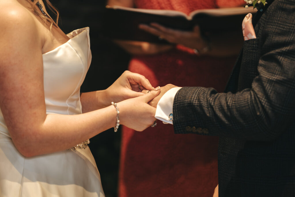 In Nottinghamshire, a photographer captures the moment a bride in a white dress places a ring on the groom's finger. The groom, in a dark suit, stands before an officiant in red. Their hands exchanging rings become the focus, symbolizing wedding vows gracefully sealed. © Aimee Lince Photography