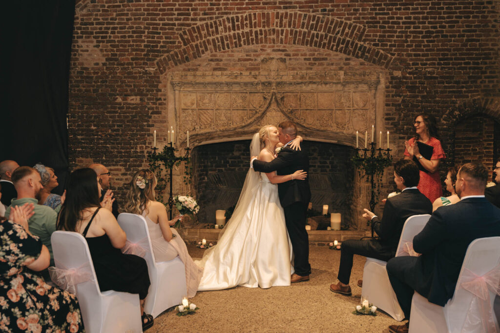 In a stunning display of wedding elegance, a bride and groom embrace by a grand stone fireplace during their Nottinghamshire ceremony. The bride's long white gown contrasts beautifully with her bouquet, as guests watch from either side. A woman in red officiates amid candles and greenery, capturing perfect photography moments. © Aimee Lince Photography