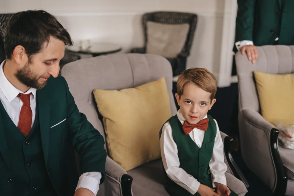 A young boy in a green vest and red bow tie sits on a gray chair, gazing at the camera with an autumnal warmth. Next to him, a man with a beard in a matching green suit looks down. Both wear white shirts. The chairs have yellow cushions, hinting at the formal charm of Ashbourne Hotel's setting. © Aimee Lince Photography - Wedding photographer in Lincolnshire, Yorkshire & Nottinghamshire