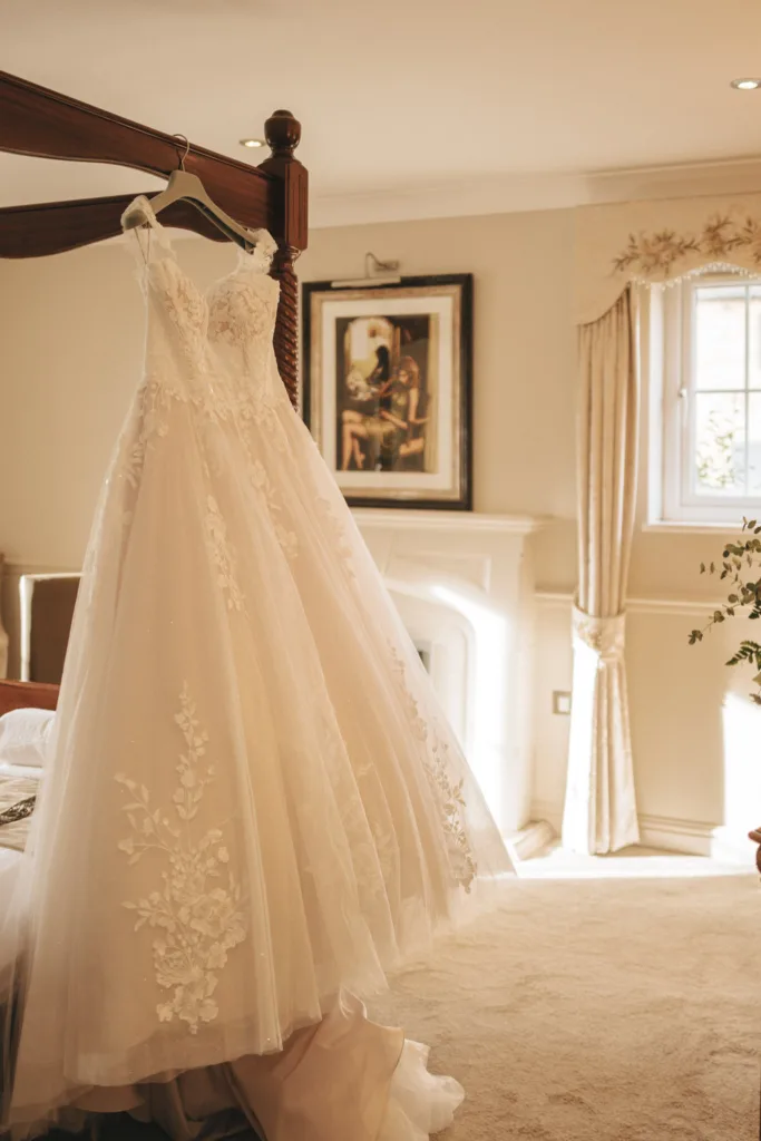 A delicate white wedding dress with lace detailing is hanging on a wooden bed frame in a sunlit room at the Ashbourne Hotel. The space boasts a fireplace, a framed painting, and a large window with light curtains. Sunlight filters softly through, casting a warm glow on the dress and beige carpet. © Aimee Lince Photography - Wedding photographer in Lincolnshire, Yorkshire & Nottinghamshire