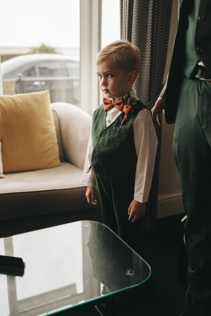 A young boy stands indoors near a window at the Ashbourne Hotel, wearing a dark green vest and pants with a white shirt. He has a bow tie and orange boutonnière, perfect for the wedding. A person's arm is beside him. The room features a beige sofa with yellow cushions and patterned curtains, ideal for photography. © Aimee Lince Photography - Wedding photographer in Lincolnshire, Yorkshire & Nottinghamshire