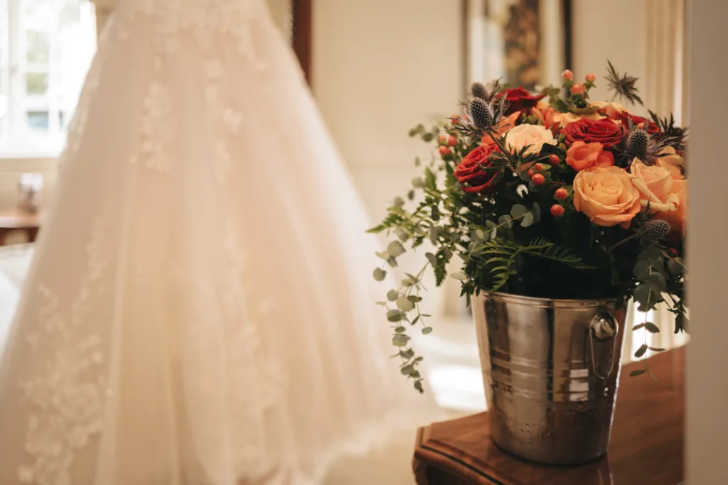 A bouquet of orange and red roses mixed with greenery sits in a metal bucket on a wooden table, capturing the essence of an autumn Ashbourne Hotel wedding. In the softly blurred background, a white lace wedding dress is visible, adding to the scene's warmth and charm. © Aimee Lince Photography - Wedding photographer in Lincolnshire, Yorkshire & Nottinghamshire