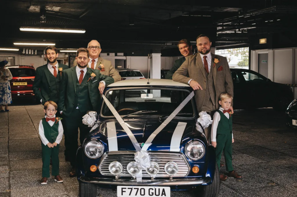 A group of seven men and two boys, dressed in green suits, brown waistcoats, and ties, stand around a classic blue Mini Cooper adorned with white ribbons and bows. In this parking garage scene, they celebrate an Ashbourne Hotel wedding with a festive flair. © Aimee Lince Photography - Wedding photographer in Lincolnshire, Yorkshire & Nottinghamshire