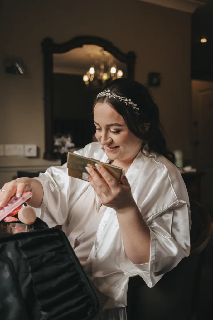 In a beautifully captured moment of photography, a woman in a white satin robe sits at a table, smiling as she holds a compact mirror and makeup item. Her hair, adorned with a jeweled headband, reflects the warm elegance reminiscent of an autumn Ashbourne Hotel wedding. © Aimee Lince Photography - Wedding photographer in Lincolnshire, Yorkshire & Nottinghamshire