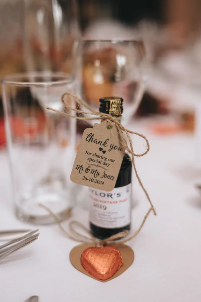 A small bottle of Taylor's Vintage Port, adorned with a rustic twine bow and a tag reading, "Thank you for sharing our special day, Mr & Mrs Jones, 26-10-2024," graces the white tablecloth at Ashbourne Hotel. Nearby, glistening glassware and heart-shaped chocolate complete the charming wedding scene. © Aimee Lince Photography - Wedding photographer in Lincolnshire, Yorkshire & Nottinghamshire
