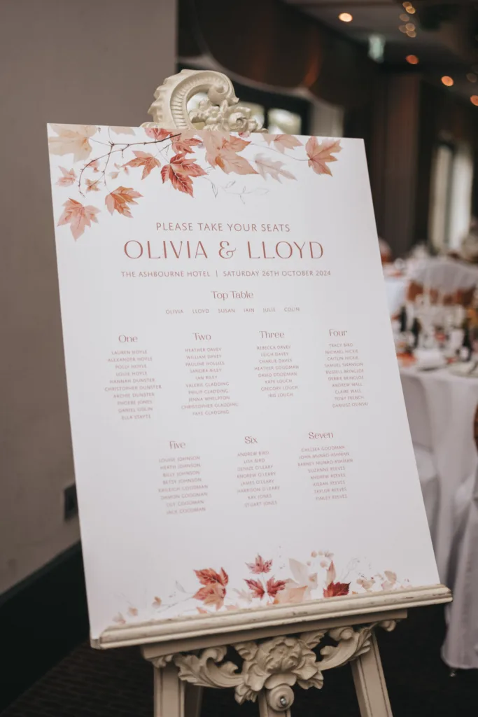 Wedding seating chart on an easel, featuring an autumn leaves design by a talented wedding photographer. It reads "Please Take Your Seats, Olivia & Lloyd, The Ashbourne Hotel, Saturday 26th October 2024." Lists tables labeled One to Seven with guest names. Decorated with red and orange leaves on a white background. © Aimee Lince Photography - Wedding photographer in Lincolnshire, Yorkshire & Nottinghamshire