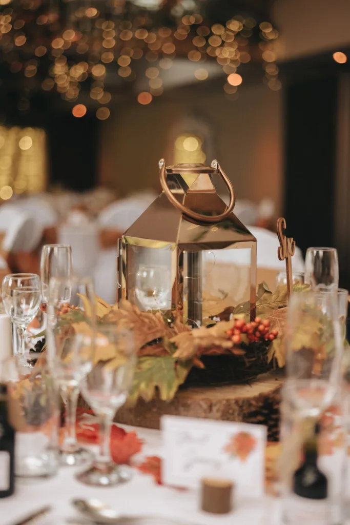 A wedding table centerpiece at the Ashbourne Hotel features a golden lantern surrounded by autumn leaves, berries, and small branches on a wooden slab. The blurred background showcases twinkling string lights and elegantly set tables with white linens and glassware, creating a warm, festive ambiance. © Aimee Lince Photography - Wedding photographer in Lincolnshire, Yorkshire & Nottinghamshire
