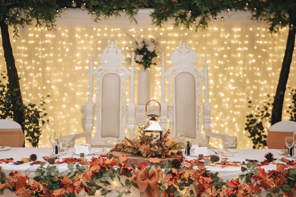 A decorated wedding reception table at the Ashbourne Hotel features a centerpiece with autumn leaves and a lantern. Behind it, two ornate white chairs sit before glowing string lights and greenery. With wine glasses and place settings meticulously arranged, it's a photographer's dream setting. © Aimee Lince Photography - Wedding photographer in Lincolnshire, Yorkshire & Nottinghamshire