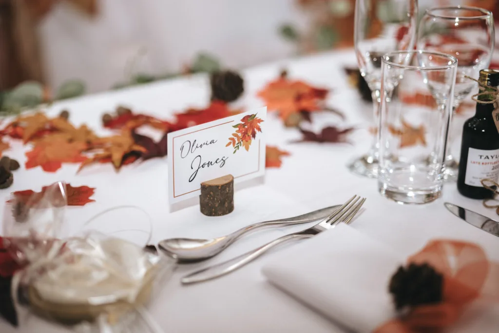 A table at the Ashbourne Hotel, set for an autumn-themed event, showcases a place card reading "Dianne Jones" in elegant script, held by a small wooden block. Surrounded by fall leaves, pine cones, glasses, cutlery, and Taylors' port wine with a white napkin nearby to complete the setting. © Aimee Lince Photography - Wedding photographer in Lincolnshire, Yorkshire & Nottinghamshire
