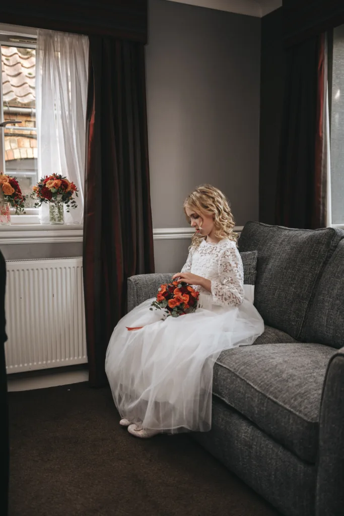 A bride in a white lace gown sits on a gray sofa at the Ashbourne Hotel, holding a bouquet of vibrant orange and red flowers. Her long blonde hair is styled in loose curls. Behind her, a window with brown curtains displays two flower arrangements echoing her autumn bouquet's colors. The room features dark carpet and light gray walls. © Aimee Lince Photography - Wedding photographer in Lincolnshire, Yorkshire & Nottinghamshire
