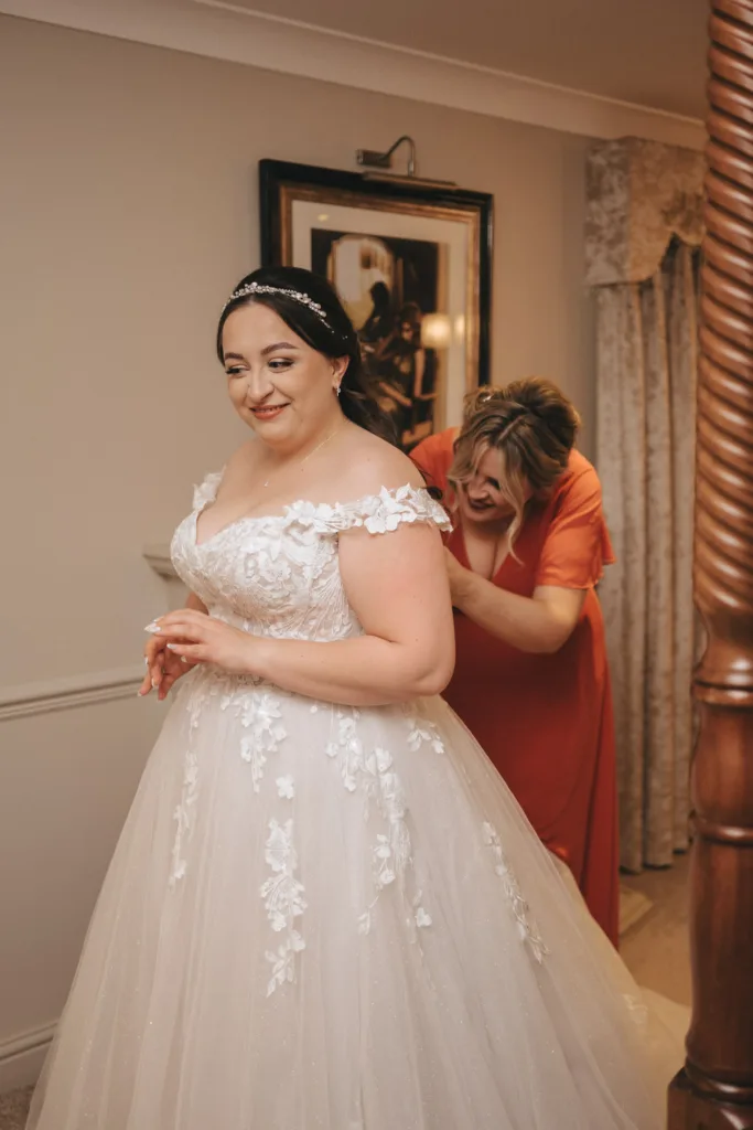 A bride in a white, off-shoulder wedding gown with floral details stands smiling while a woman in a red dress adjusts her dress. The scene captures the essence of autumn wedding photography, set in a warmly lit room with framed painting and draped curtains in the background. © Aimee Lince Photography - Wedding photographer in Lincolnshire, Yorkshire & Nottinghamshire