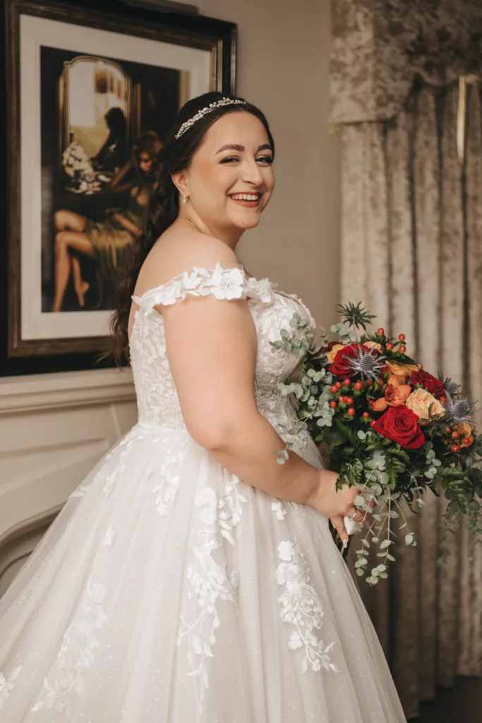 The bride, at the Ashbourne Hotel wedding, in an off-shoulder lace-embellished white gown smiles while holding a vibrant bouquet of red and orange flowers with greenery. Her hair is elegantly styled with a headband, set against a backdrop of a framed painting and elegant drapes. © Aimee Lince Photography - Wedding photographer in Lincolnshire, Yorkshire & Nottinghamshire