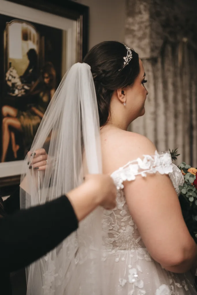 In the soft, neutral light of an Ashbourne hotel suite, a bride stands in an off-shoulder lace gown and sheer veil. As a person adjusts her dress, she clutches an autumn bouquet. A framed painting with two figures hangs nearby, capturing a moment as if through timeless photography. © Aimee Lince Photography - Wedding photographer in Lincolnshire, Yorkshire & Nottinghamshire