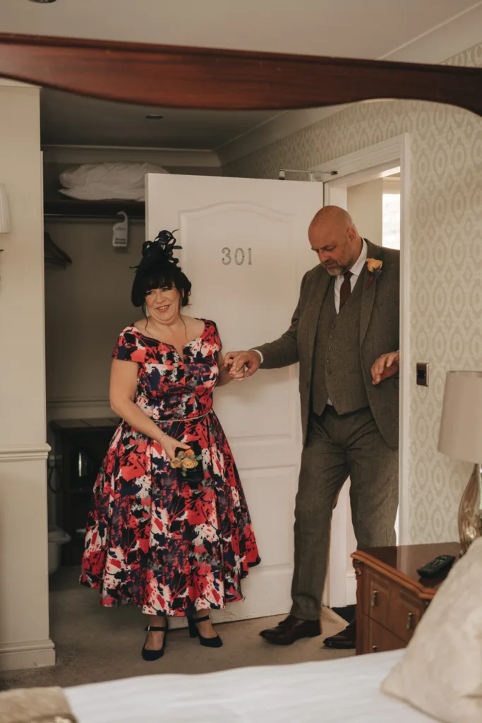 A woman in a vibrant floral dress, wearing a black hat, smiles as she holds a bouquet and stands in a doorway marked "301" at the Ashbourne Hotel. A man in a brown suit with a peach boutonniere gently holds her hand. The room is warmly lit, with a wooden dresser and lamp visible, capturing perfect wedding photography. © Aimee Lince Photography - Wedding photographer in Lincolnshire, Yorkshire & Nottinghamshire