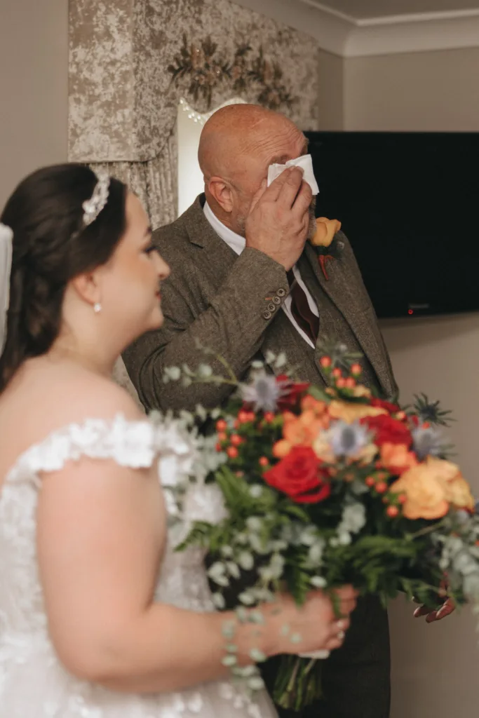A bride in a white lace dress holds a colorful bouquet at The Ashbourne Hotel, while an older man in a brown suit wipes his eyes with a tissue. The room, decorated with neutral-colored walls and a flat-screen TV, exudes an emotional atmosphere before the autumn wedding ceremony. © Aimee Lince Photography - Wedding photographer in Lincolnshire, Yorkshire & Nottinghamshire