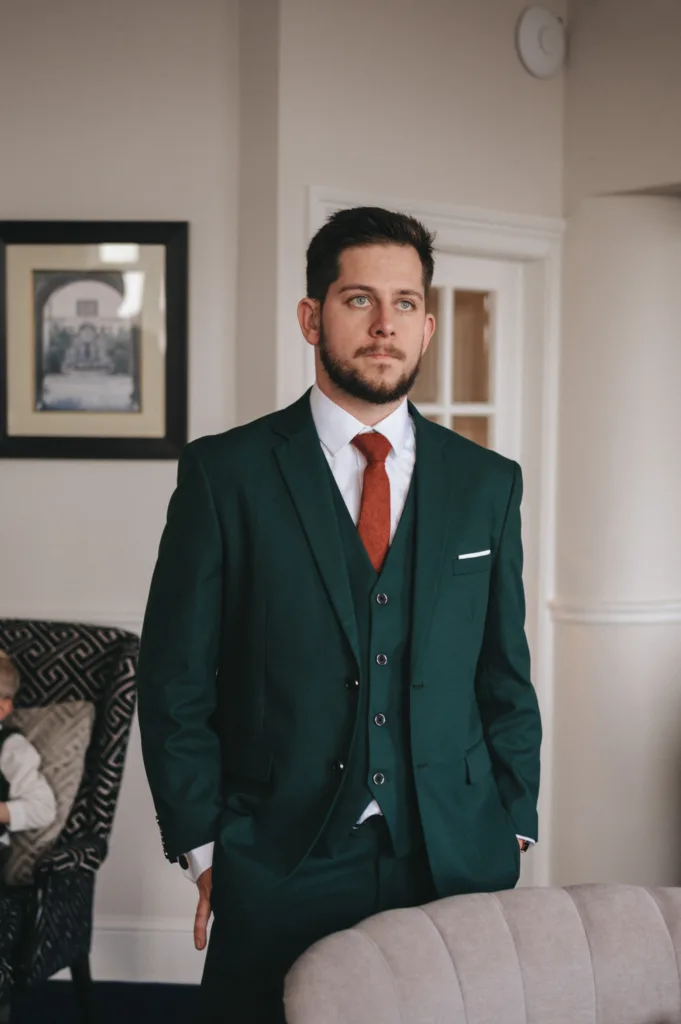 A man with a beard stands in a room, wearing a dark green three-piece suit, a white dress shirt, and a red tie—perfect for wedding photography. His hands are in his pockets as he gazes thoughtfully to the side. A framed picture hangs on the wall, and a patterned chair is visible in the background. © Aimee Lince Photography - Wedding photographer in Lincolnshire, Yorkshire & Nottinghamshire
