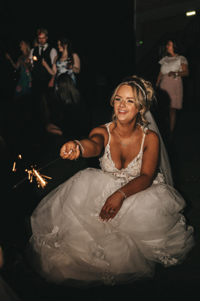 A bride in a white wedding dress sits on the grass at night at Oaklands Hall, holding a sparkler. Her hair is styled elegantly with a veil. In the background, guests are gathered, with some standing and others mingling. The joyful scene glows warmly as captured by wedding photography enthusiasts. © Aimee Lince Photography