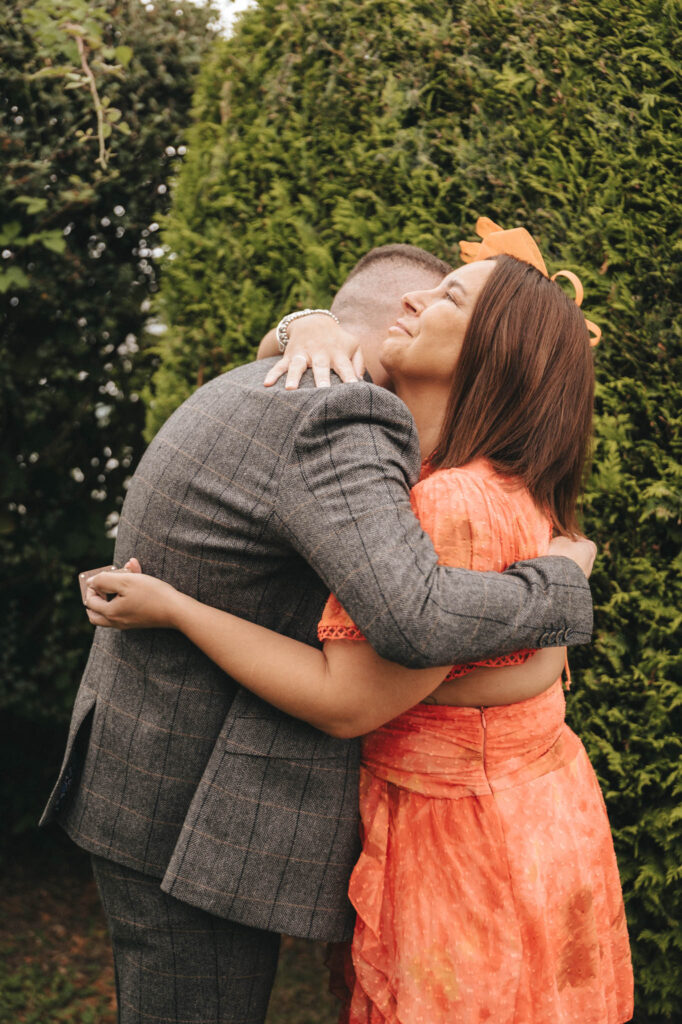 A man in a gray plaid suit and a woman in an orange floral dress embrace warmly outdoors at The Oaklands. Her long brown hair is adorned with a flower headpiece, perfect for their wedding photos amidst the lush green foliage that creates a serene natural backdrop. © Aimee Lince Photography