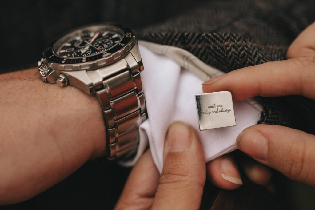 Close-up of a person adjusting a cufflink with an engraved message, "with you today and always," perfect for wedding photos at Oaklands Hall Hotel. The cufflink is paired with a silver watch on the left wrist, with the hand holding a white shirt cuff partially covered by a brown herringbone jacket sleeve. © Aimee Lince Photography