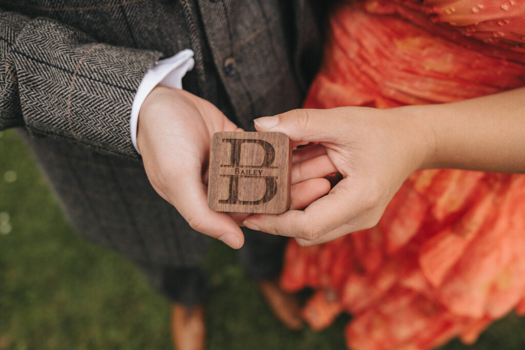 A person in a brown tweed jacket holds a wooden block with the letter "B" and the word "Bailey" engraved on it, reminiscent of keepsakes found at Oaklands Hall weddings. Another person in an orange dress reaches to touch the block. They are standing on grass, with focus on their hands and the wooden block. © Aimee Lince Photography