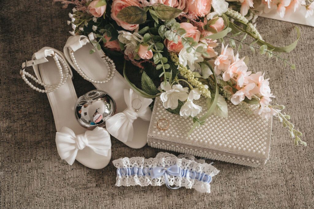 A flat lay of bridal accessories for wedding photography features white shoes with ankle straps and bows, a pearl-embellished clutch, a bouquet of pink and white flowers, a lace garter with blue ribbon, a silver ring, and a perfume bottle. Perfectly arranged on a textured gray surface at Oaklands Hall. © Aimee Lince Photography