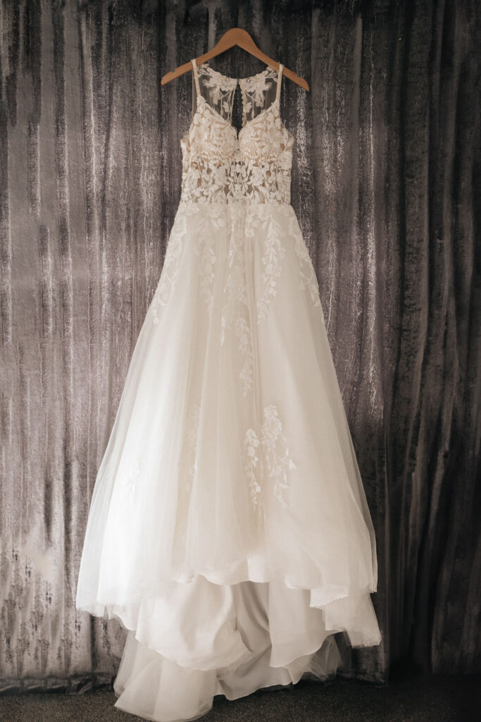 A white wedding dress hangs on a wooden hanger against a textured gray curtain, ready for the perfect wedding photos. The sleeveless lace bodice features floral embroidery, and the flowing tulle skirt with a subtle train creates an elegant and romantic look, ideal for ceremonies at Oaklands Hall. © Aimee Lince Photography