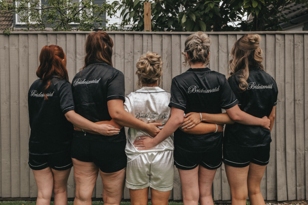 Five women stand with their backs to the camera, arms around each other at Oaklands Hall Hotel. Four in matching black outfits have "Bridesmaid" embroidered, while one in white has "Bride" on her back. This charming wedding photo captures them against a wooden fence with greenery above. © Aimee Lince Photography
