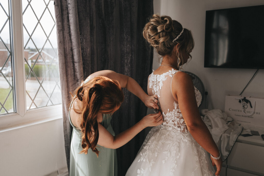 A bride in a detailed lace wedding dress stands as another woman with long red hair helps fasten the back. They are near a window with diamond-patterned panes and a dark curtain at Oaklands Hall. A flat-screen TV and a box labeled "Coates Couture" are visible in the background, capturing perfect wedding photography moments. © Aimee Lince Photography